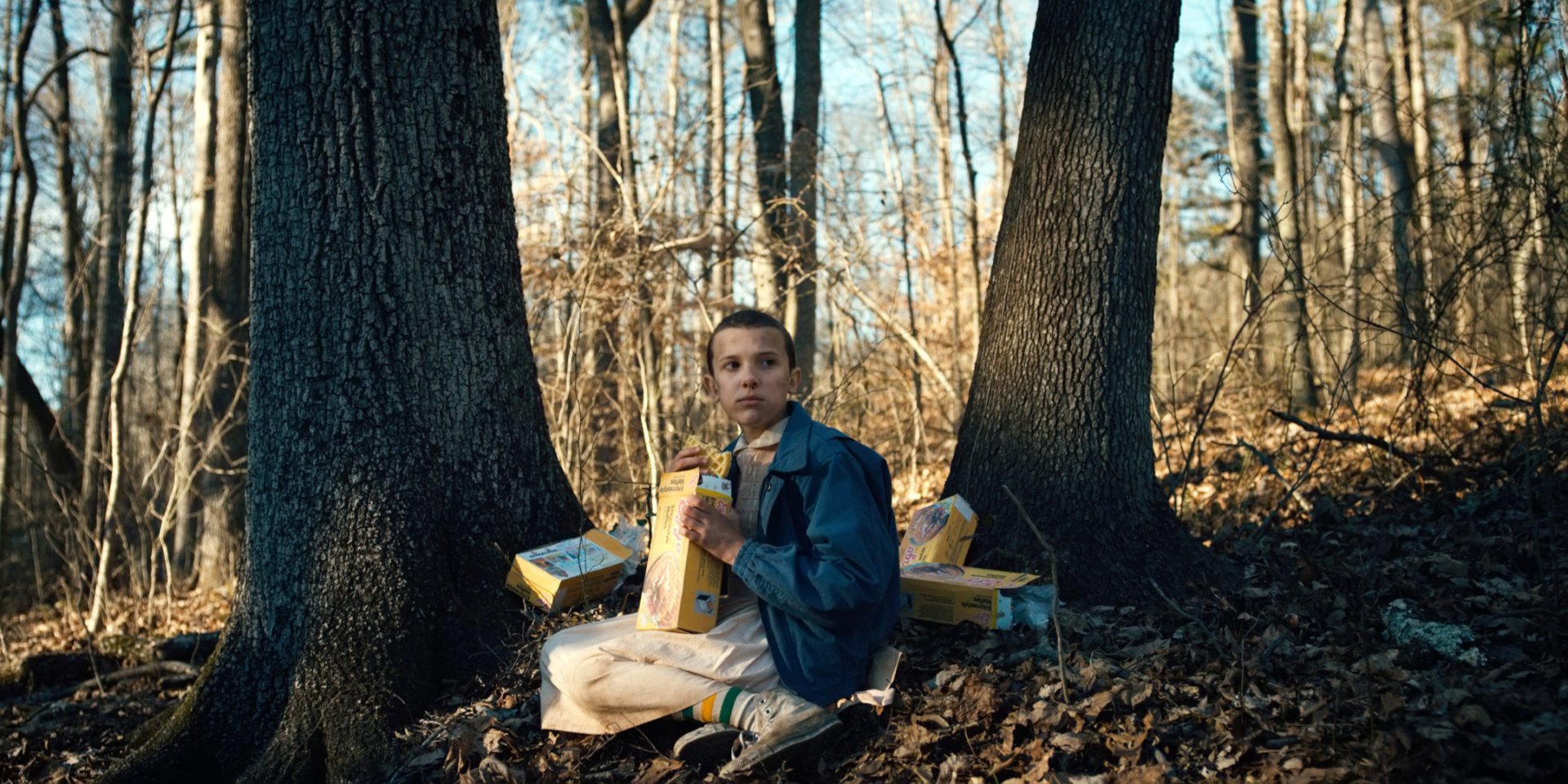 Eleven from Stranger Things, played by Millie Bobby Brown, sits in a forest clearing surrounded by Eggo waffle boxes in this HD PC desktop wallpaper.
