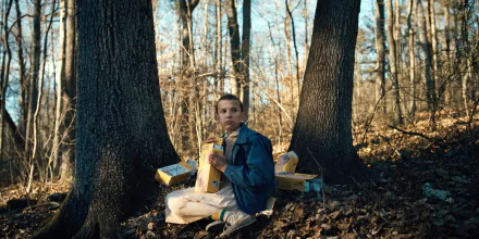 Eleven from Stranger Things, played by Millie Bobby Brown, sits in a forest clearing surrounded by Eggo waffle boxes in this HD PC desktop wallpaper.