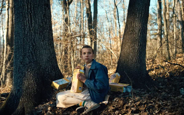 Eleven from Stranger Things, played by Millie Bobby Brown, sits in a forest clearing surrounded by Eggo waffle boxes in this HD PC desktop wallpaper.
