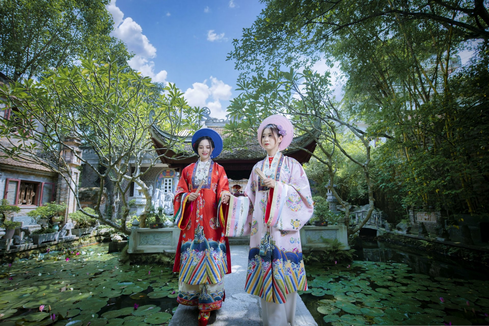 8K Ultra HD PC desktop wallpaper: two Asian women in colorful traditional dress standing amid lotus pads before a temple, framed by trees and a bright blue sky.