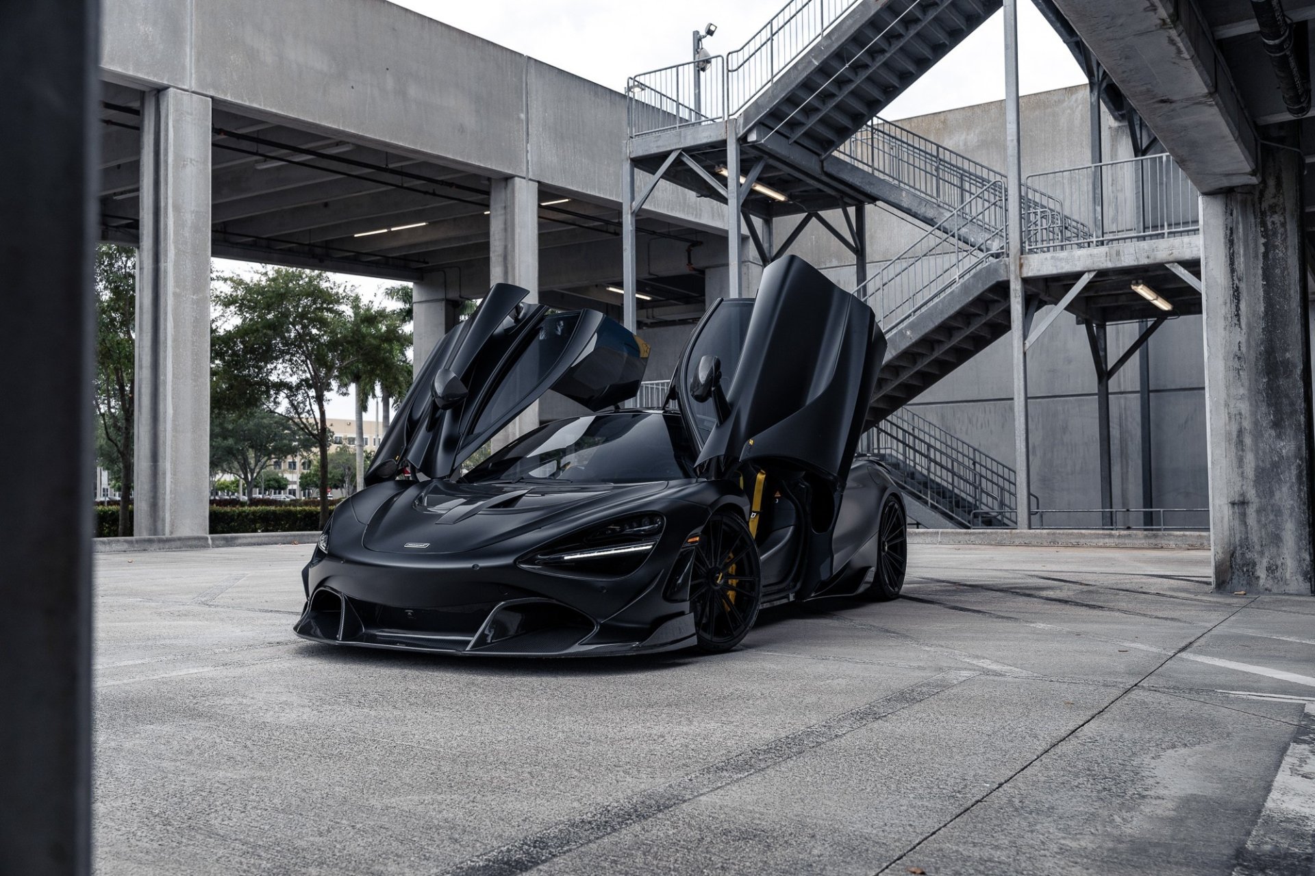 HD PC desktop wallpaper featuring a sleek black McLaren 720S with its butterfly doors open in an urban concrete setting.