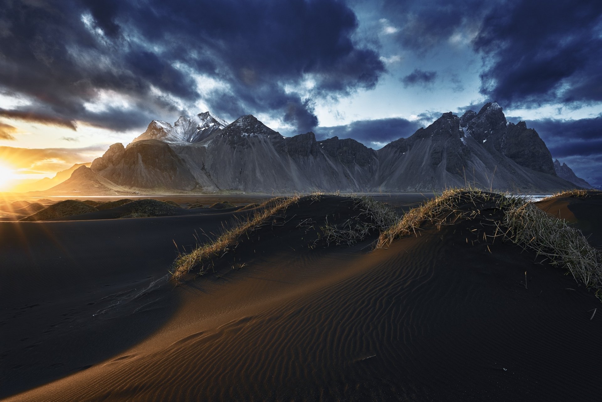 HD PC desktop wallpaper of Vestrahorn: black sand dunes and grassy hummocks lead to jagged mountains beneath a dramatic, stormy sky as sunlight breaks the horizon.