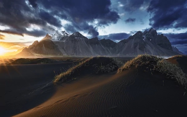 HD PC desktop wallpaper of Vestrahorn: black sand dunes and grassy hummocks lead to jagged mountains beneath a dramatic, stormy sky as sunlight breaks the horizon.