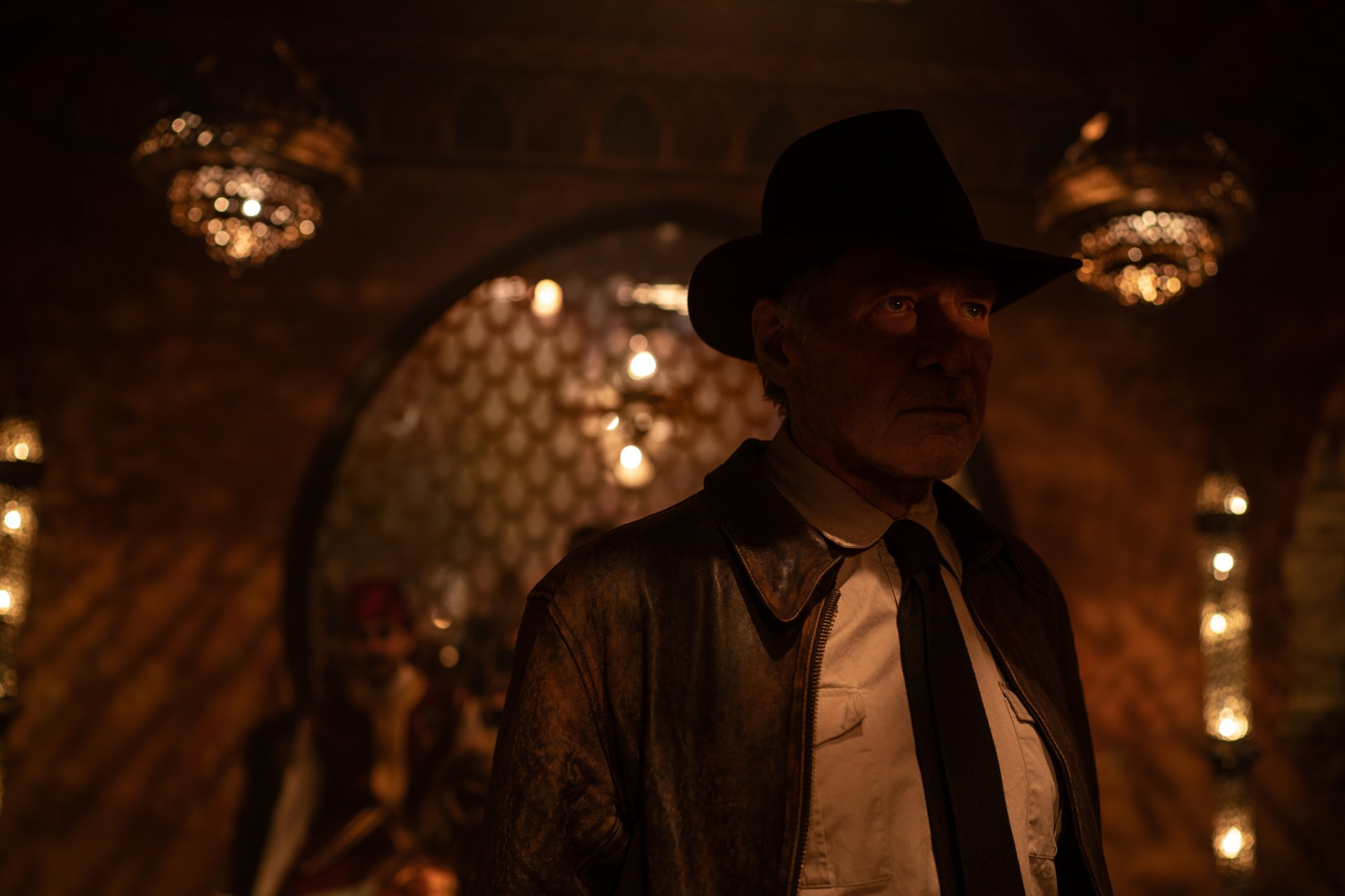 Brooding adventurer in fedora and leather jacket in a dim, ornate room, cinematic poster for Indiana Jones and the Dial of Destiny — 4K Ultra HD PC desktop wallpaper.
