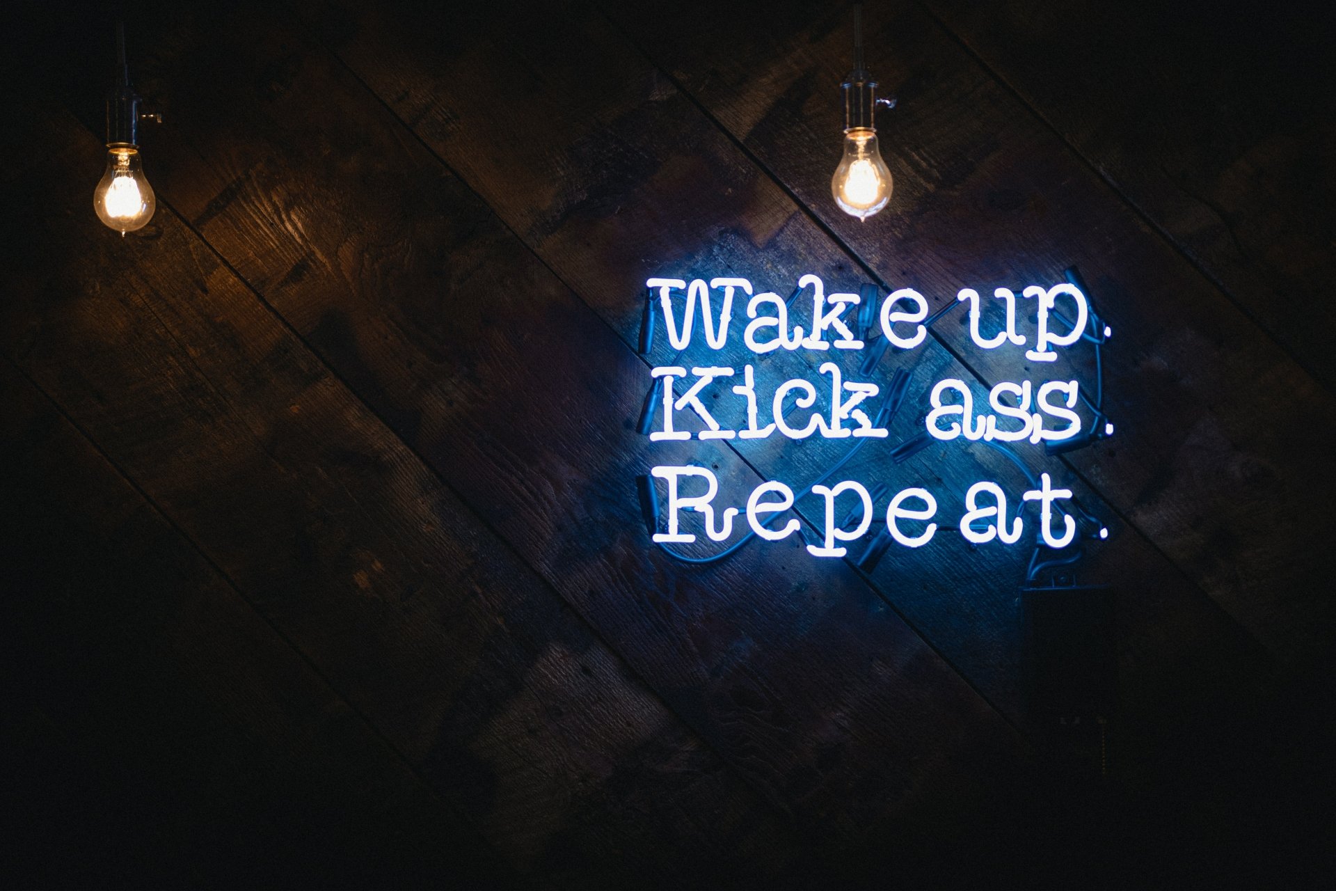 Motivational blue neon sign reading Wake up. Kick ass. Repeat. with hanging bulbs on a dark textured wall — artistic neon 5K Ultra HD PC desktop wallpaper/background.