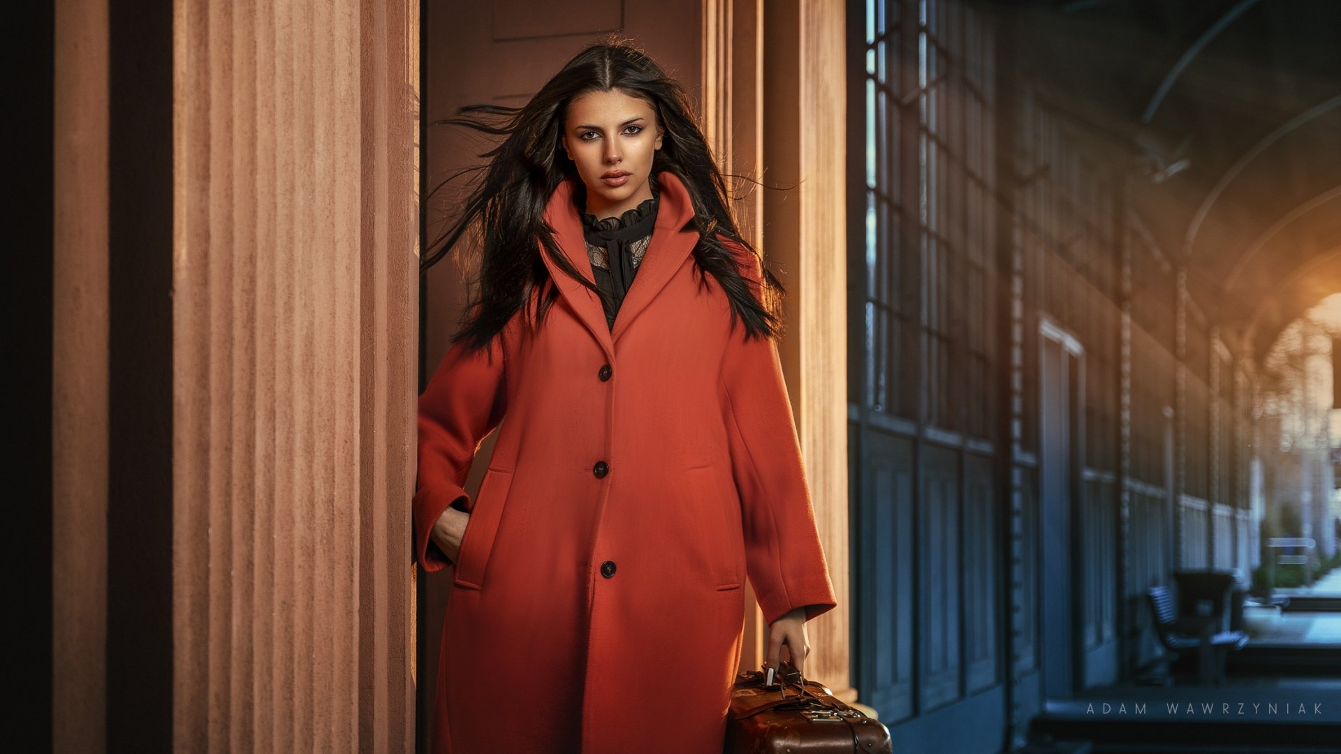 HD PC desktop wallpaper of a woman model in a red coat walking through an elegant columned corridor, carrying a vintage suitcase against a moody, cinematic background.