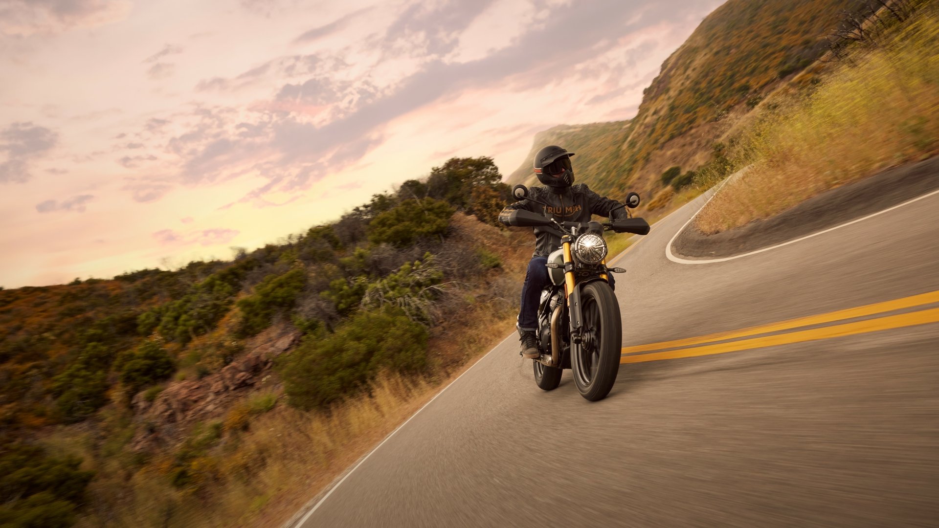 A Triumph Scrambler 400 X motorcycle navigates a winding road surrounded by lush hills, set against a vibrant sunset backdrop. HD desktop wallpaper showcasing adventure.