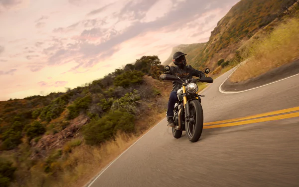 A Triumph Scrambler 400 X motorcycle navigates a winding road surrounded by lush hills, set against a vibrant sunset backdrop. HD desktop wallpaper showcasing adventure.