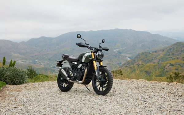 A Triumph Scrambler 400 X parked on gravel with a stunning mountain backdrop, showcasing its rugged design in this HD desktop wallpaper.