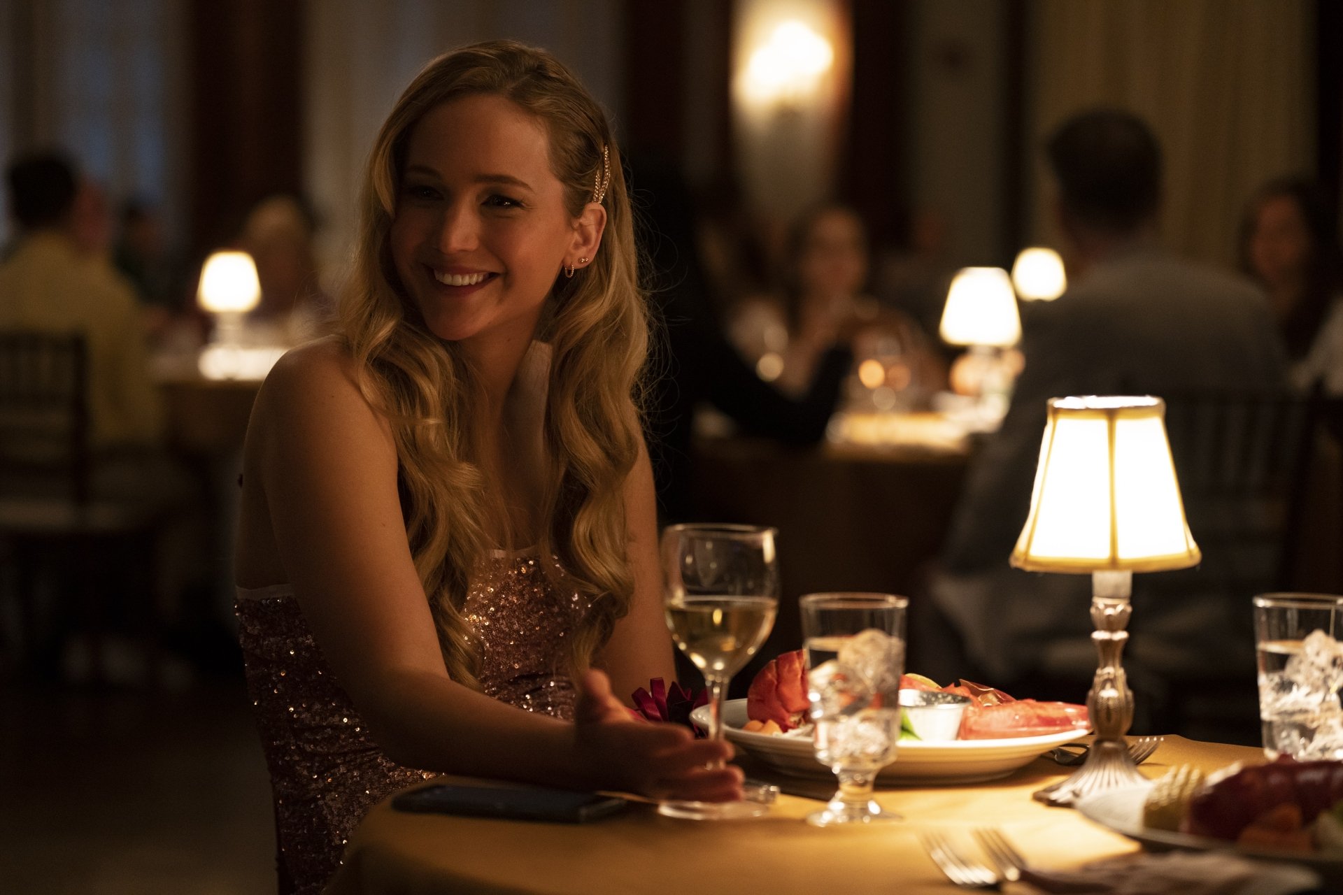 A scene from the movie No Hard Feelings, featuring a woman in a glamorous dress smiling at a dimly lit restaurant table, surrounded by elegantly set surroundings. High-definition wallpaper.
