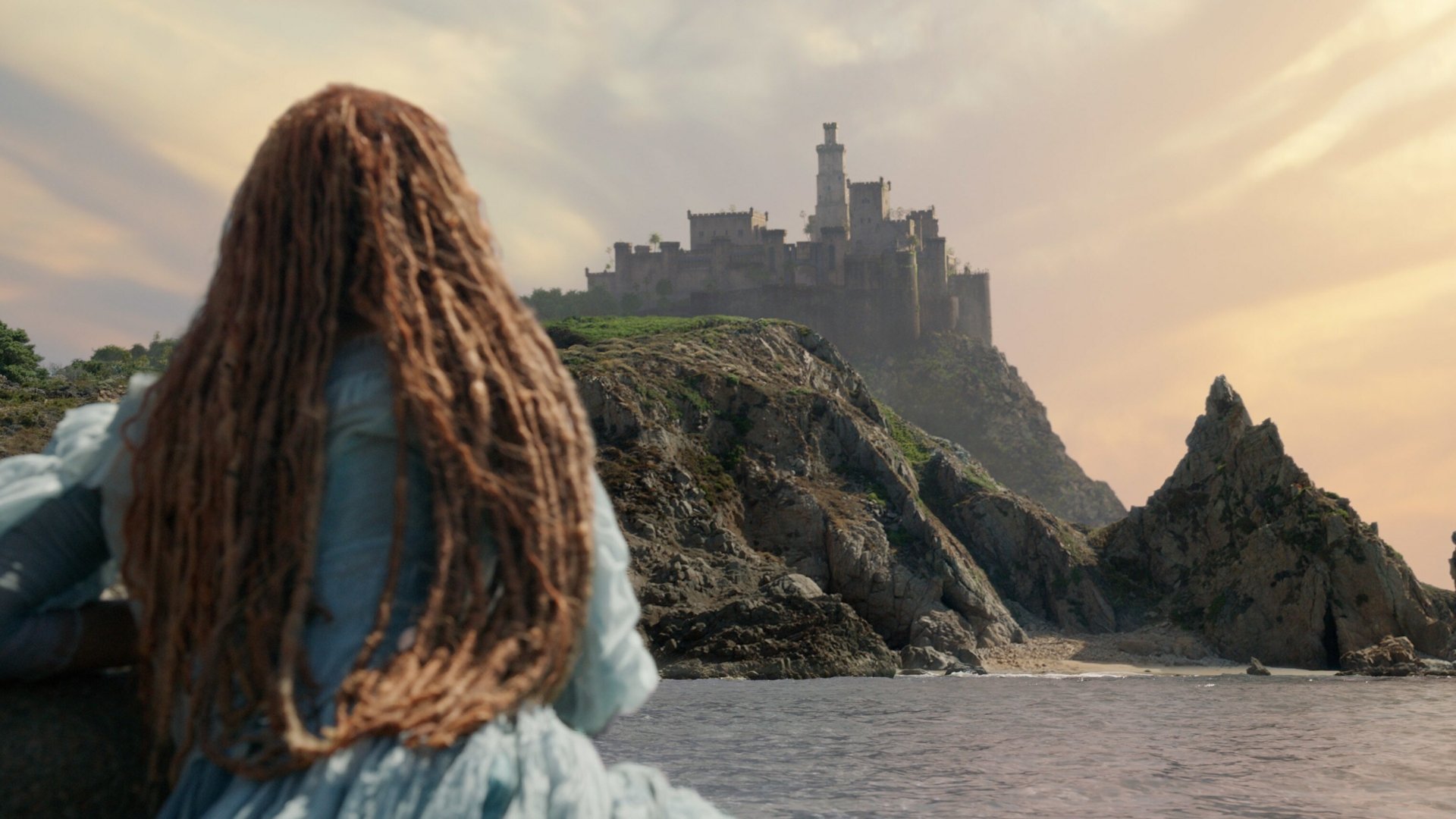 A scene from *The Little Mermaid* (2023) featuring a young woman with long, dreadlocked hair gazing at a distant castle on a rocky shore against a beautiful sunset backdrop.
