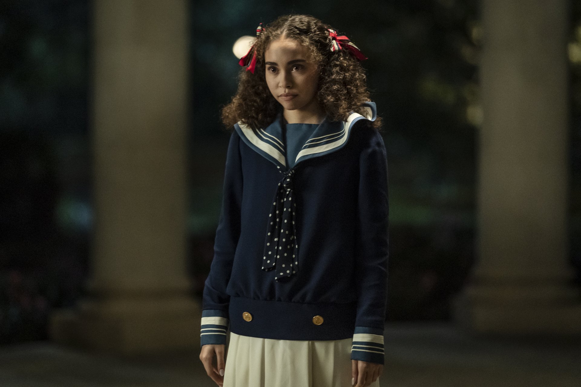 HD wallpaper featuring a character from Interview with the Vampire series portrayed by actress Bailey Bass, standing with a solemn expression in a vintage-style school uniform against a dimly lit, atmospheric backdrop.