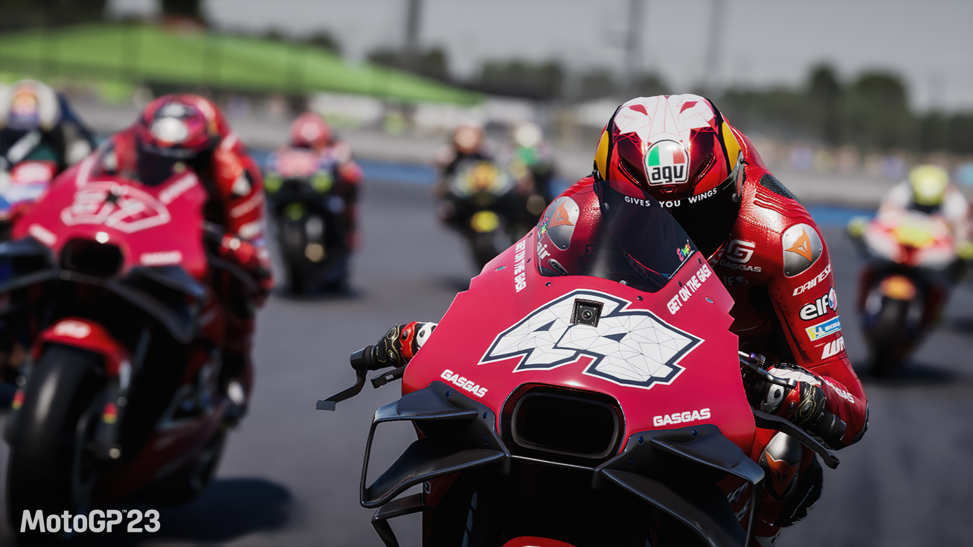 HD desktop wallpaper from the video game MotoGP 23 featuring a detailed scene of a motorcycle racer in the lead during a race, followed closely by competitors.