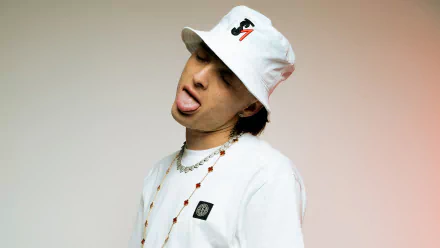 A vibrant HD desktop wallpaper featuring Peso Pluma, a Mexican musician, playfully posing in a white hat and t-shirt, showcasing his unique style and charisma.