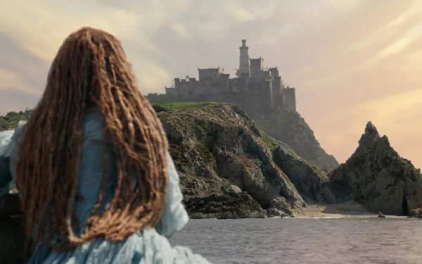 A scene from *The Little Mermaid* (2023) featuring a young woman with long, dreadlocked hair gazing at a distant castle on a rocky shore against a beautiful sunset backdrop.