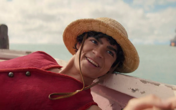 HD wallpaper of a live-action portrayal of Monkey D. Luffy from One Piece, smiling on a boat with a straw hat.
