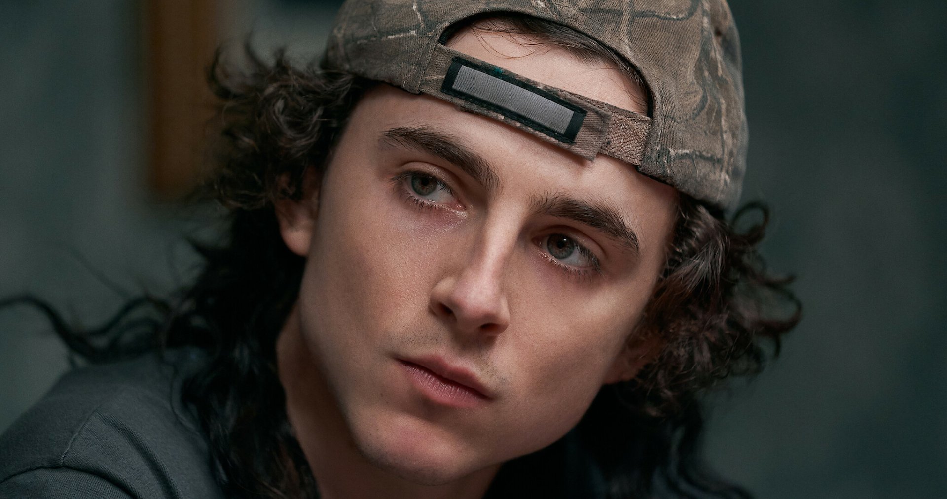 High-definition desktop wallpaper featuring a close-up of a young man with curly hair wearing a camouflage cap, providing a cinematic and moody background vibe.