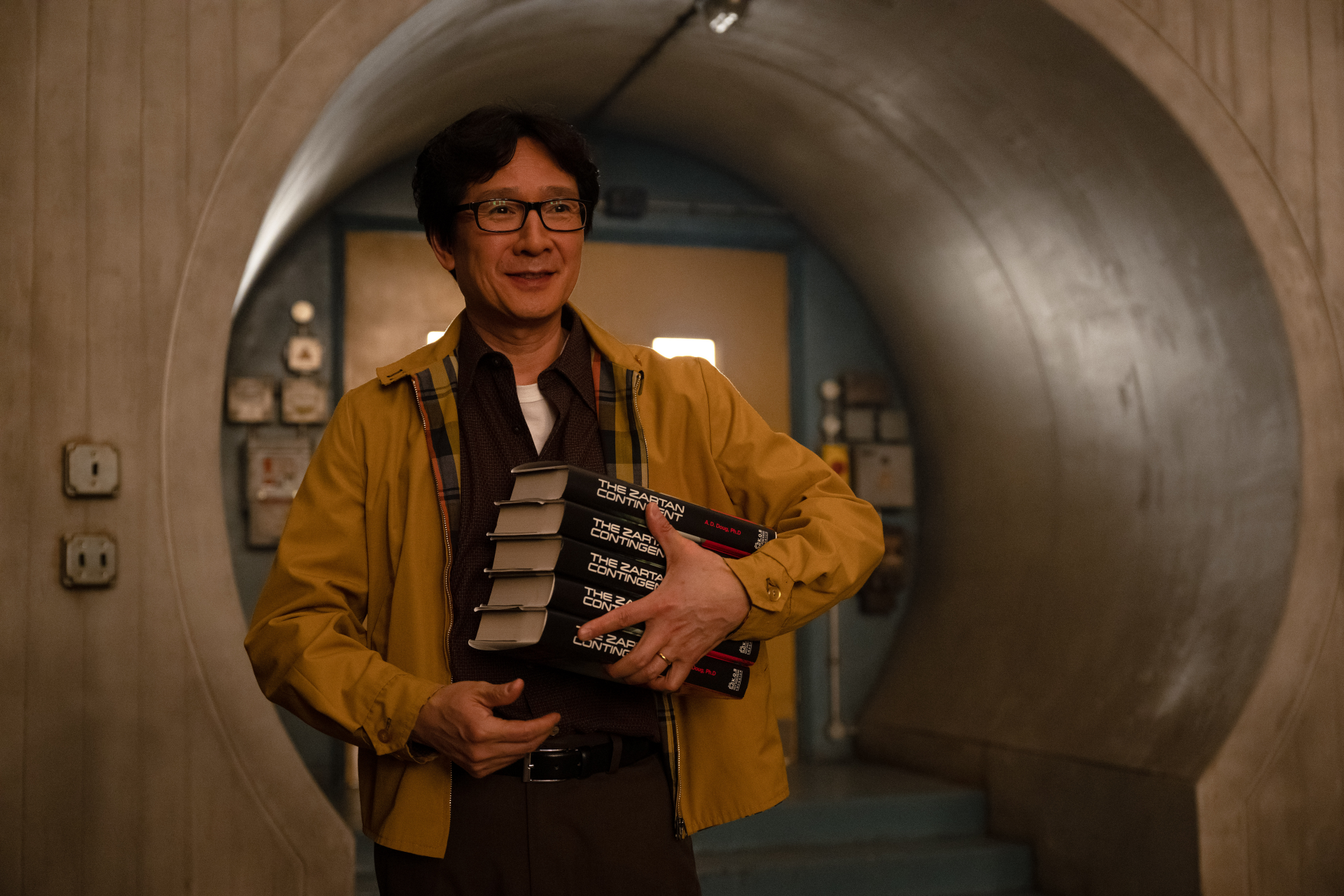 Ke Huy Quan smiles while holding books in a scene from the TV show Loki. The image serves as a vibrant HD desktop wallpaper and background.