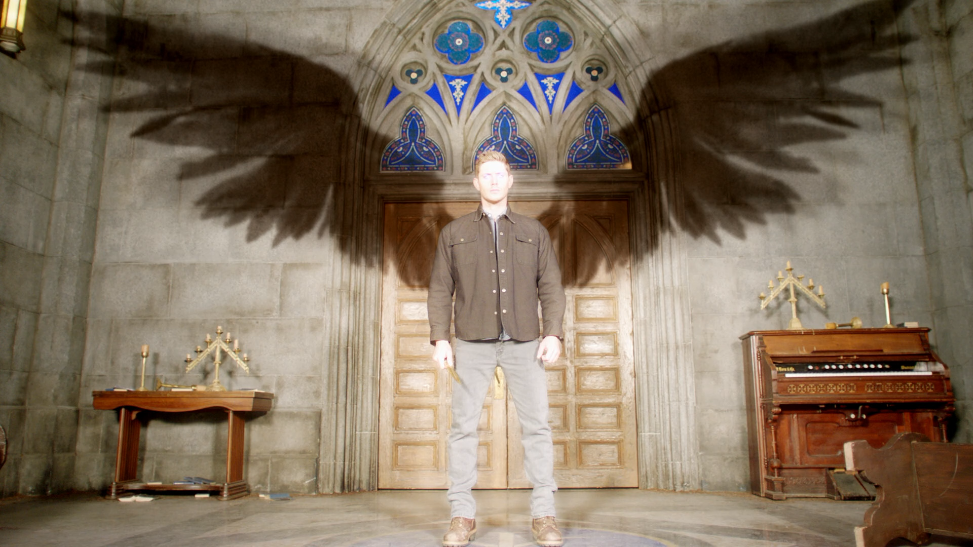 HD desktop wallpaper featuring Jensen Ackles from the TV show Supernatural, standing in a dimly lit room with large shadowed angel wings on the wall behind him.