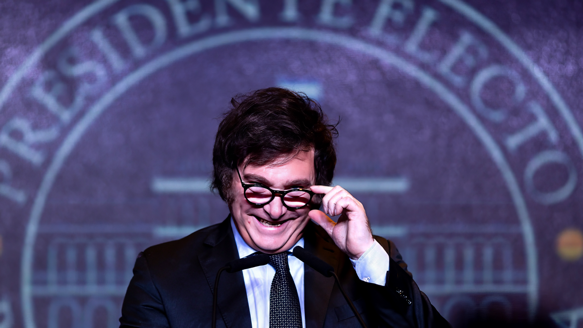Javier Milei smiles as he adjusts his glasses, set against a backdrop featuring the words Presidente Electo, making for a striking HD desktop wallpaper and background.