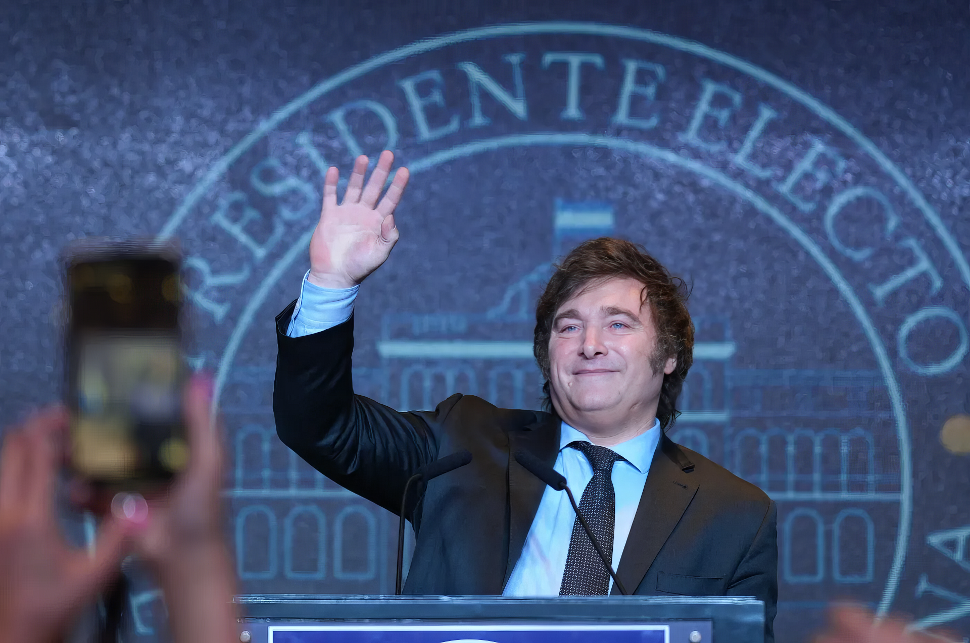 HD desktop wallpaper featuring a smiling individual waving from behind a podium with the words 'PRESIDENTE ELECTO' in the backdrop, as a foreground smartphone captures the moment.