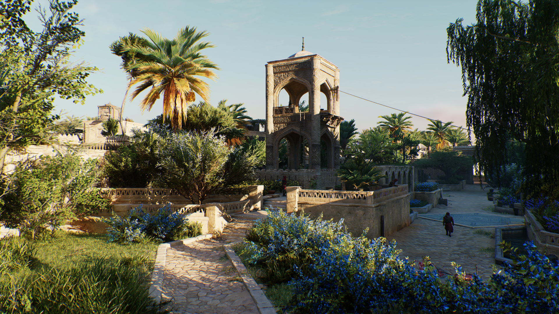 Assassin's Creed Mirage video game HD PC desktop wallpaper: sunlit Middle Eastern garden courtyard with stone pavilion, palms, blue flowers and a lone figure on a dusty path.