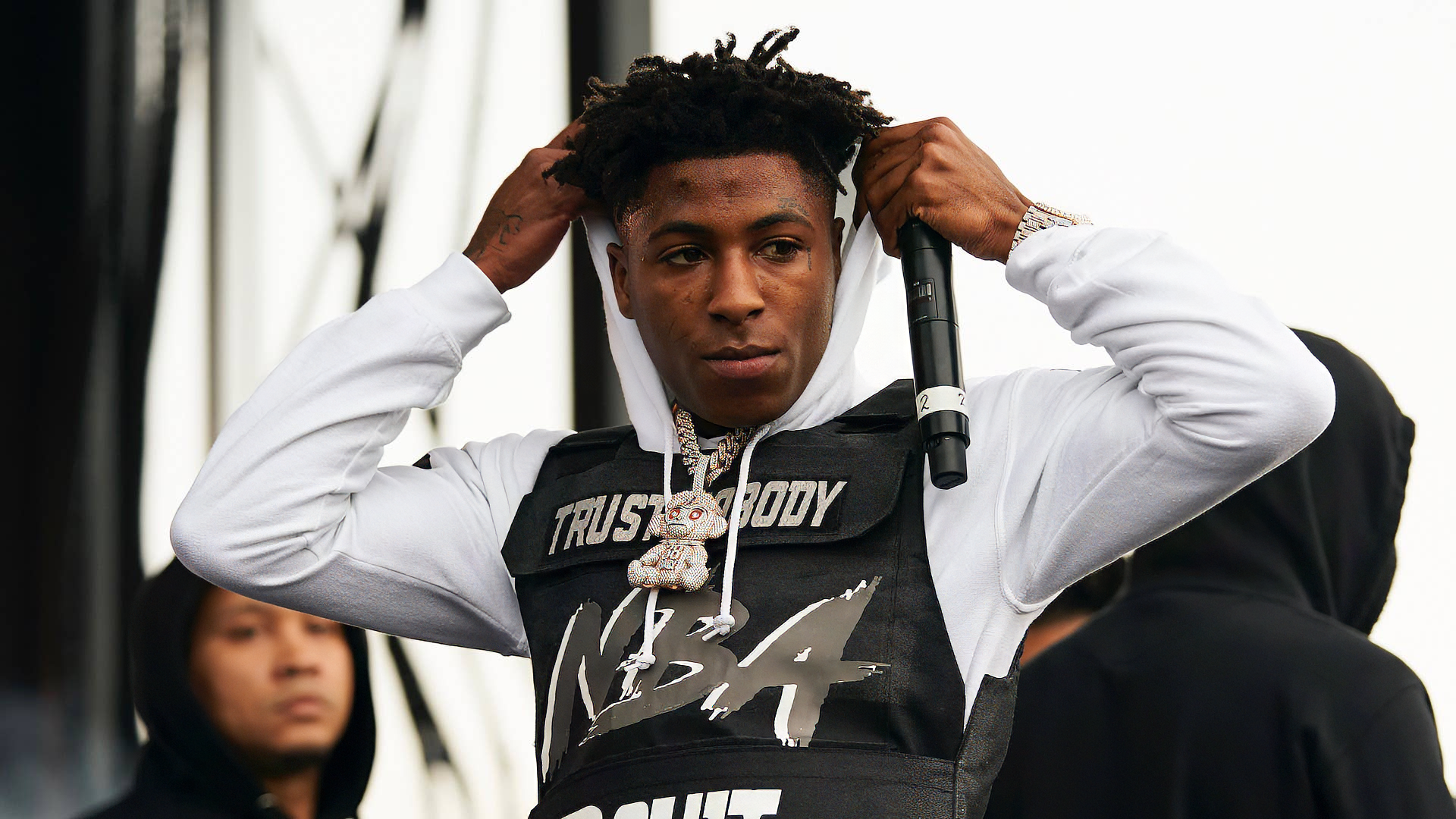 YoungBoy Never Broke Again adjusts his hooded sweatshirt while holding a microphone on stage, showcasing a dynamic energy in this HD desktop wallpaper background.