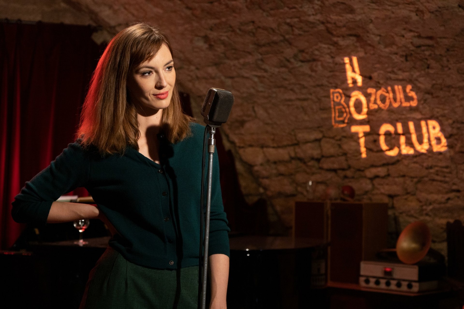 A young woman stands confidently at a microphone in a cozy club setting, with warm lighting and a stone wall backdrop, capturing a moment from the TV show Monsieur Spade.