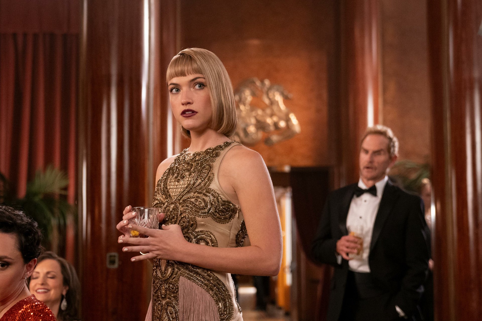 A stylish scene from the TV show Death and Other Details, featuring a woman in a glamorous dress, holding a drink, with a man in a tuxedo in the background, set in an elegant interior.