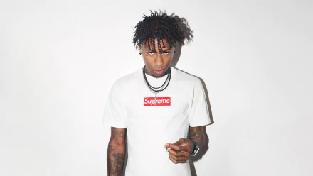 Music artist standing against a white background wearing a Supreme shirt, suitable as an HD desktop wallpaper and background for fans of YoungBoy Never Broke Again.