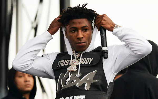 YoungBoy Never Broke Again adjusts his hooded sweatshirt while holding a microphone on stage, showcasing a dynamic energy in this HD desktop wallpaper background.