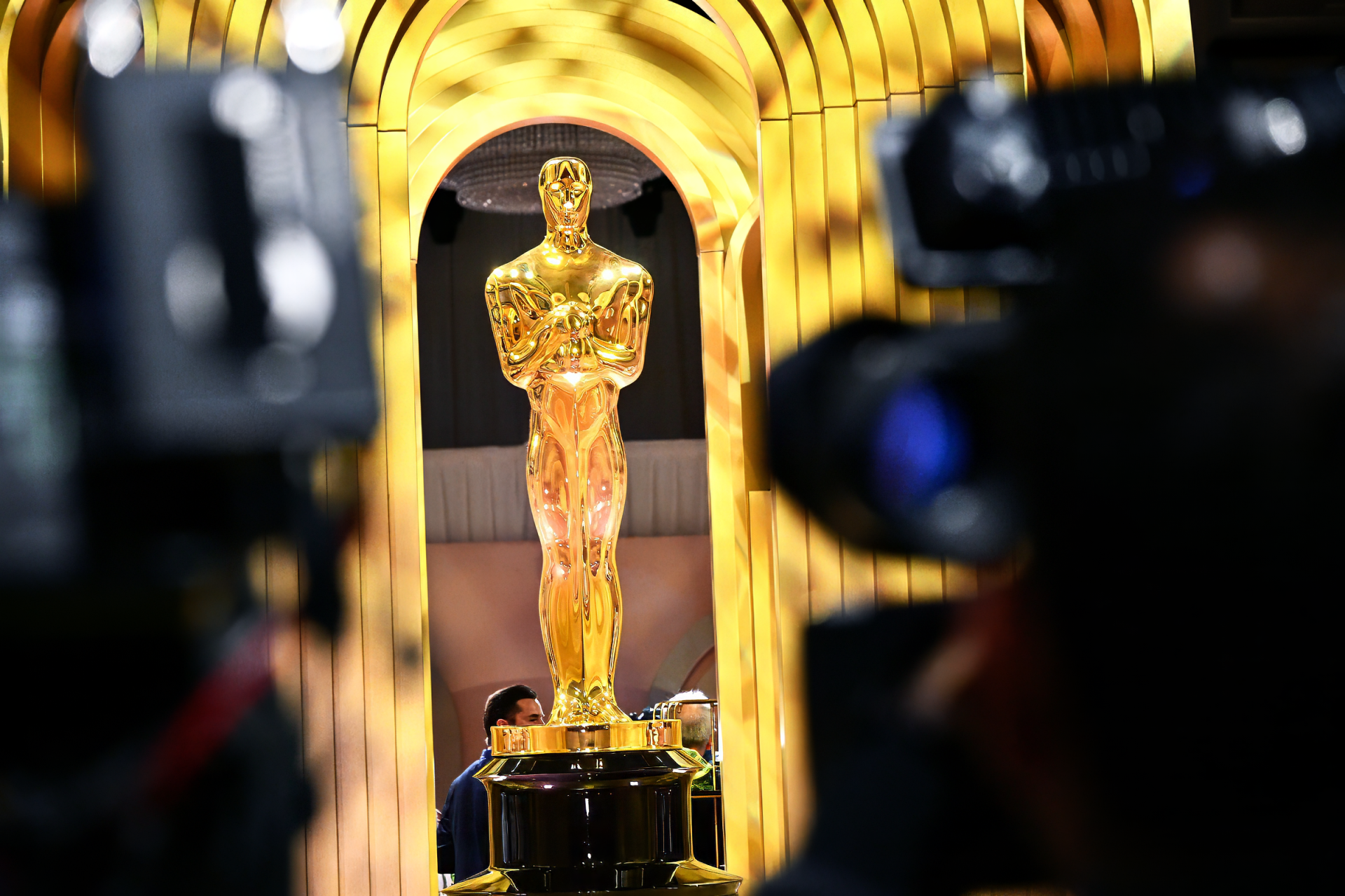 Oscar statuette in sharp focus with a golden blurred background and camera silhouette – HD Academy Awards themed desktop wallpaper.