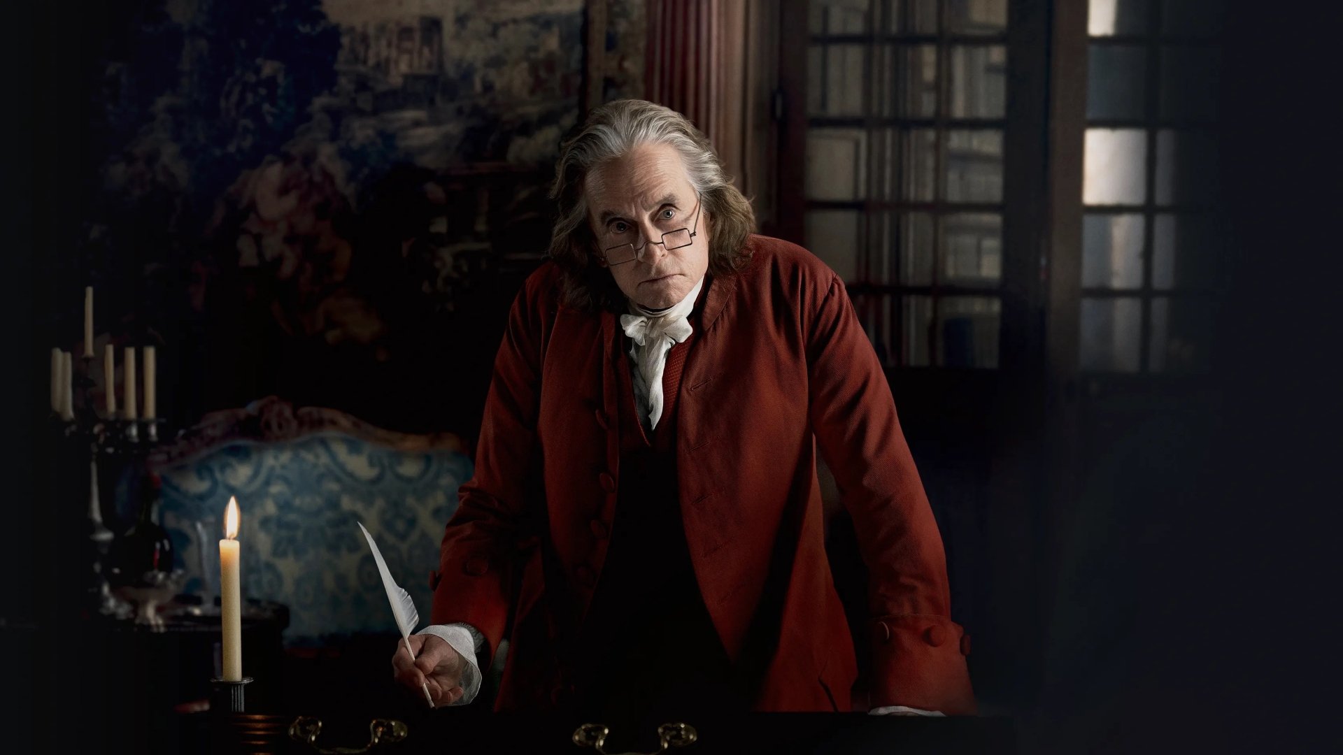 A HD desktop wallpaper from the TV show Franklin featuring a character, portrayed by an actor, dressed in historical attire with a red coat, standing pensively at a desk with a quill in hand, illuminated by candlelight.