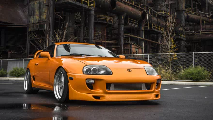 A striking orange Toyota Supra parked in front of an industrial backdrop, showcasing its sleek design and custom wheels, making an eye-catching HD desktop wallpaper.