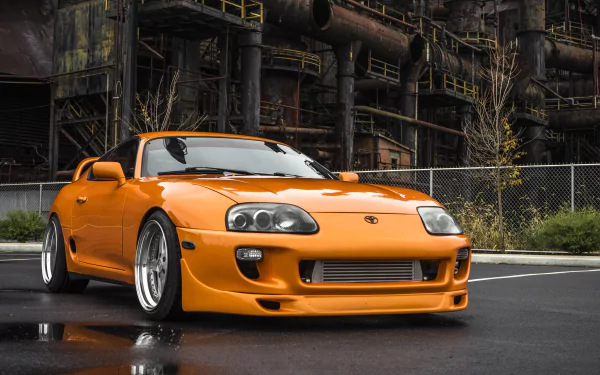 A striking orange Toyota Supra parked in front of an industrial backdrop, showcasing its sleek design and custom wheels, making an eye-catching HD desktop wallpaper.