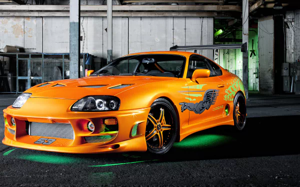 HD wallpaper of a vibrant orange Toyota Supra with stylish decals, showcased in an industrial setting.