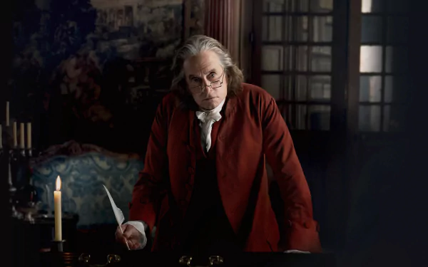 A HD desktop wallpaper from the TV show Franklin featuring a character, portrayed by an actor, dressed in historical attire with a red coat, standing pensively at a desk with a quill in hand, illuminated by candlelight.