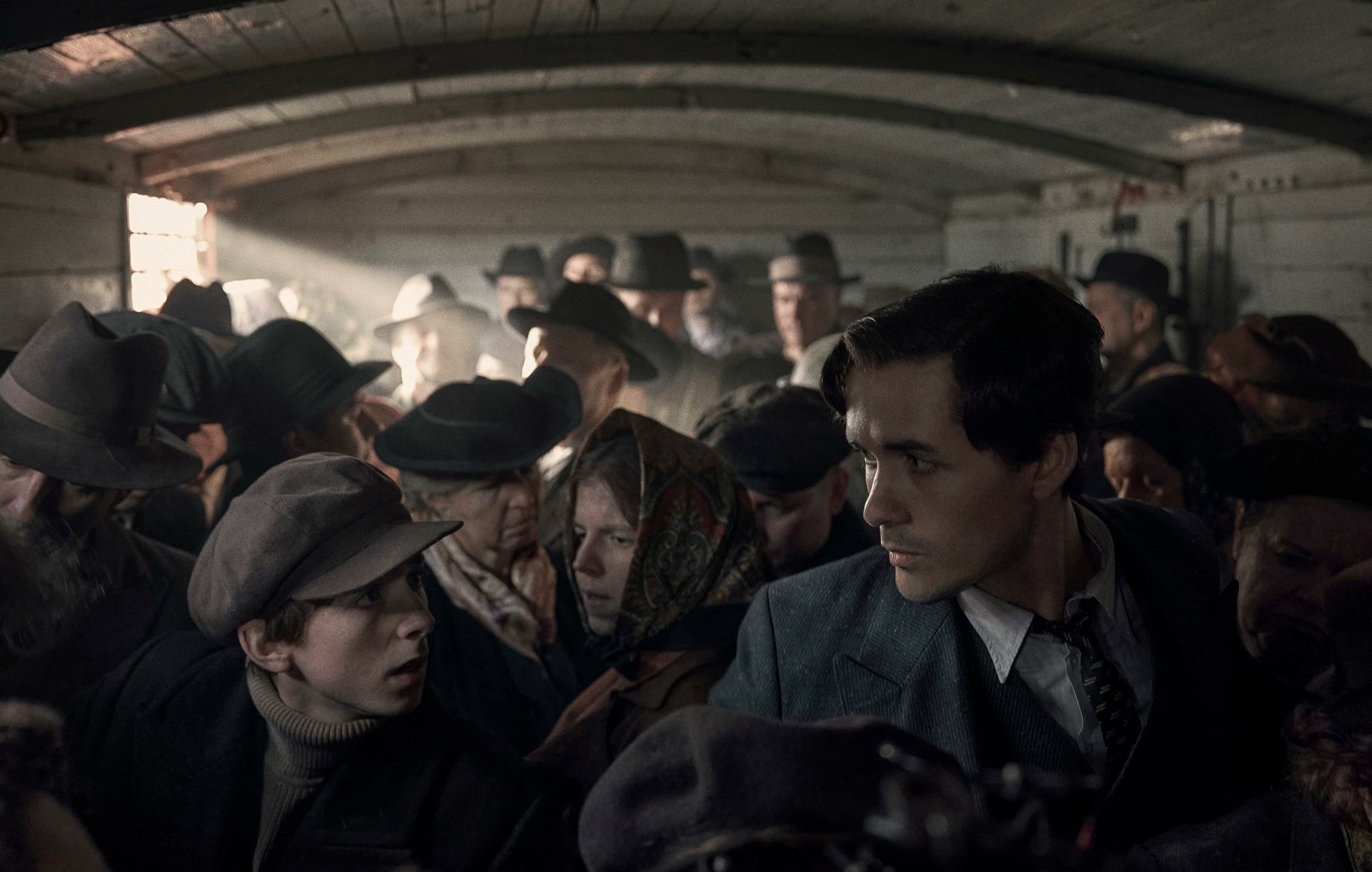HD desktop wallpaper from the TV show The Tattooist of Auschwitz, depicting a crowded scene with a thoughtful young man looking ahead.