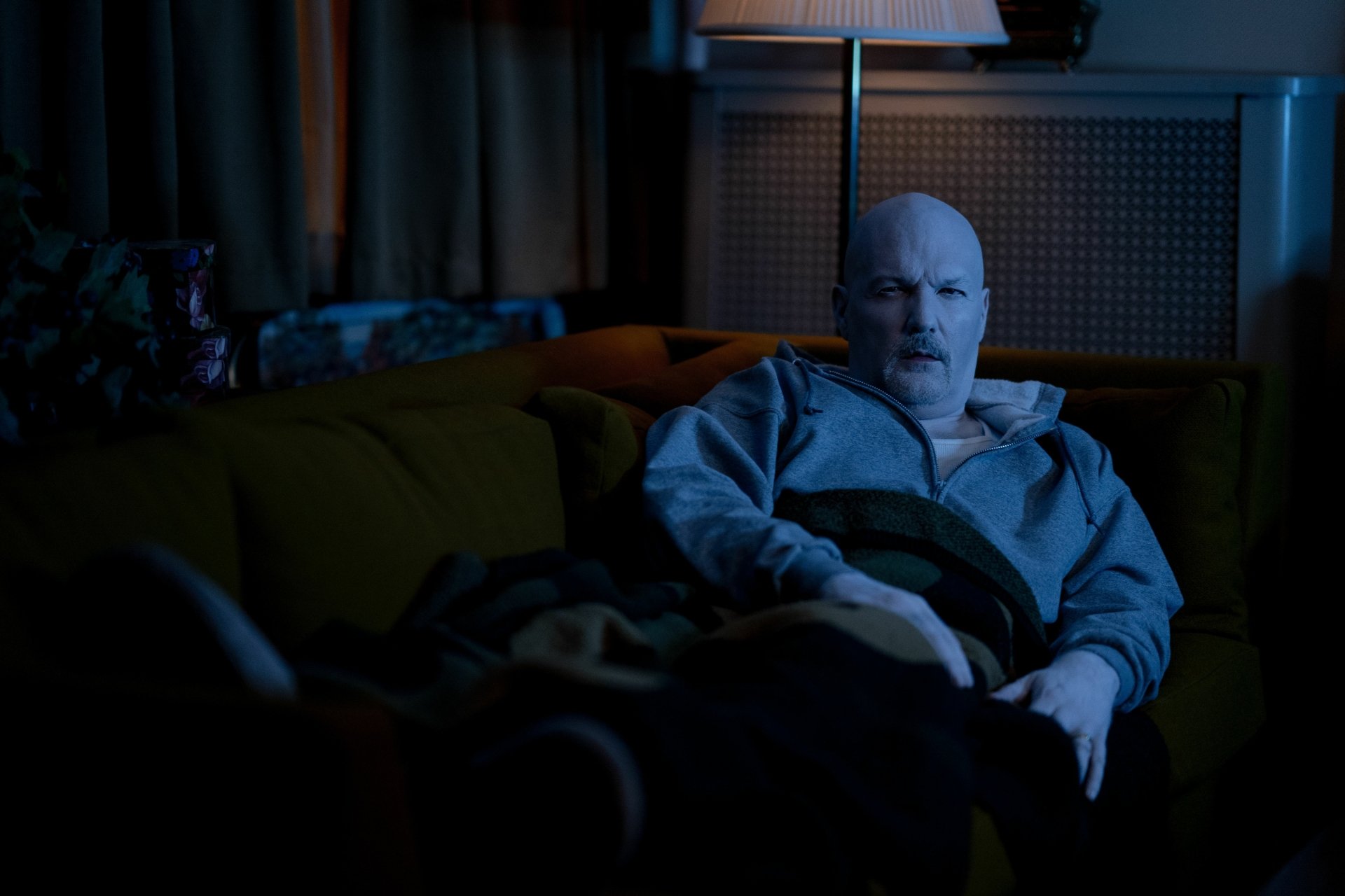 HD desktop wallpaper featuring a scene from the movie I Saw the TV Glow, with a man sitting on a couch, bathed in the blue glow of the television in a dimly lit room.