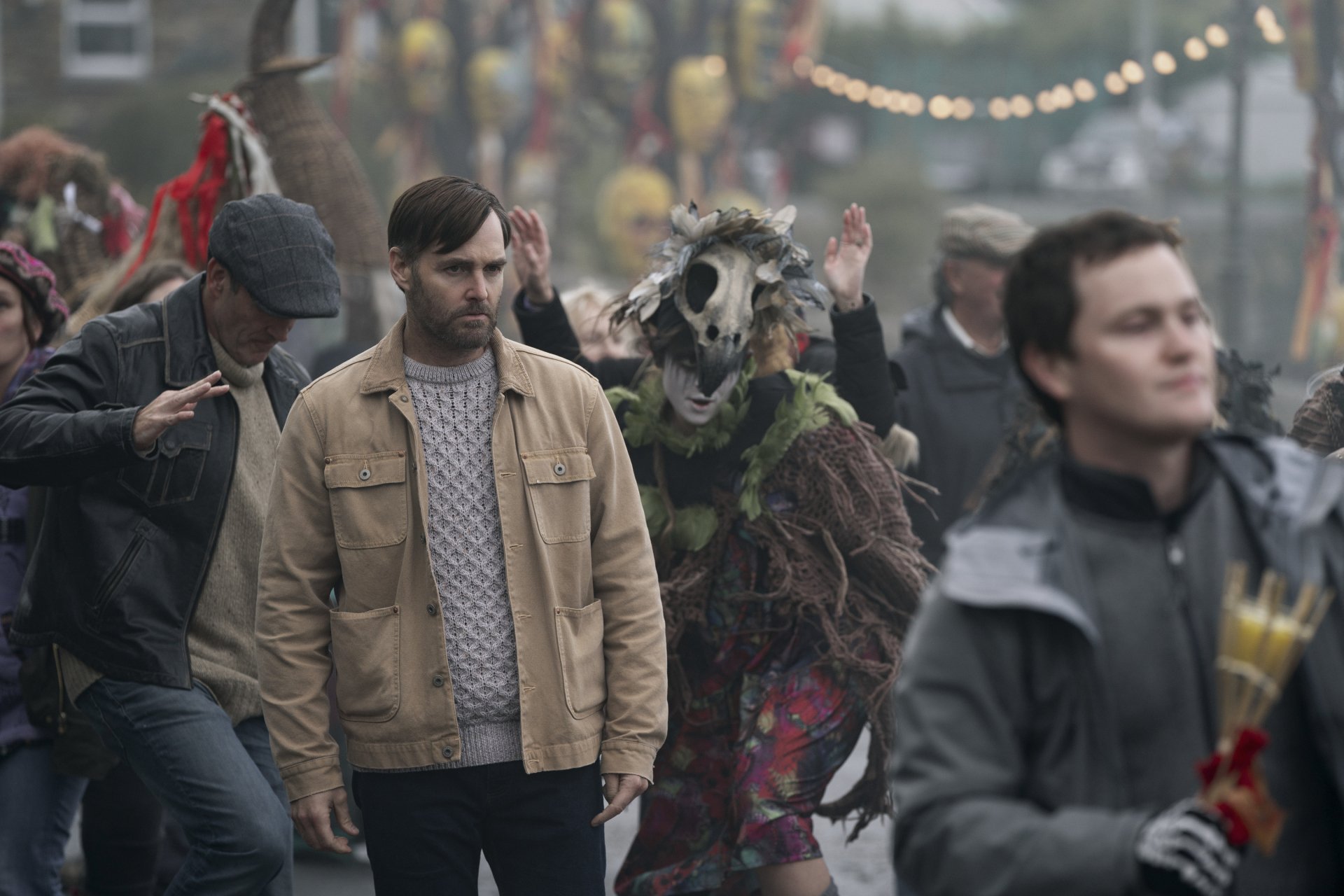 HD desktop wallpaper from the TV show Bodkin featuring a man in a beige jacket amidst a festive, chaotic street scene with masked and costumed characters.