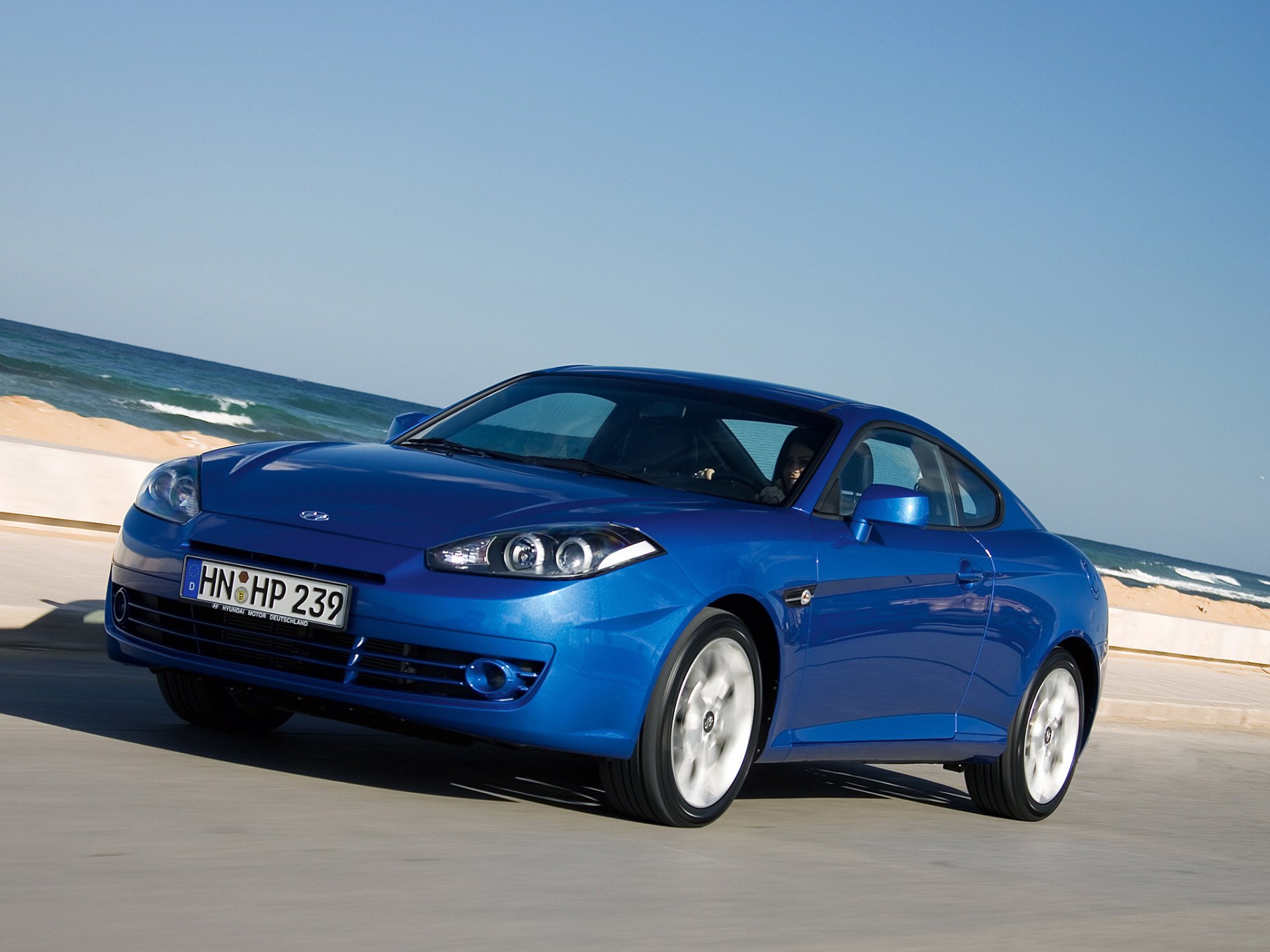 HD desktop wallpaper of a blue Hyundai Coupe parked by the seaside with clear skies in the background.