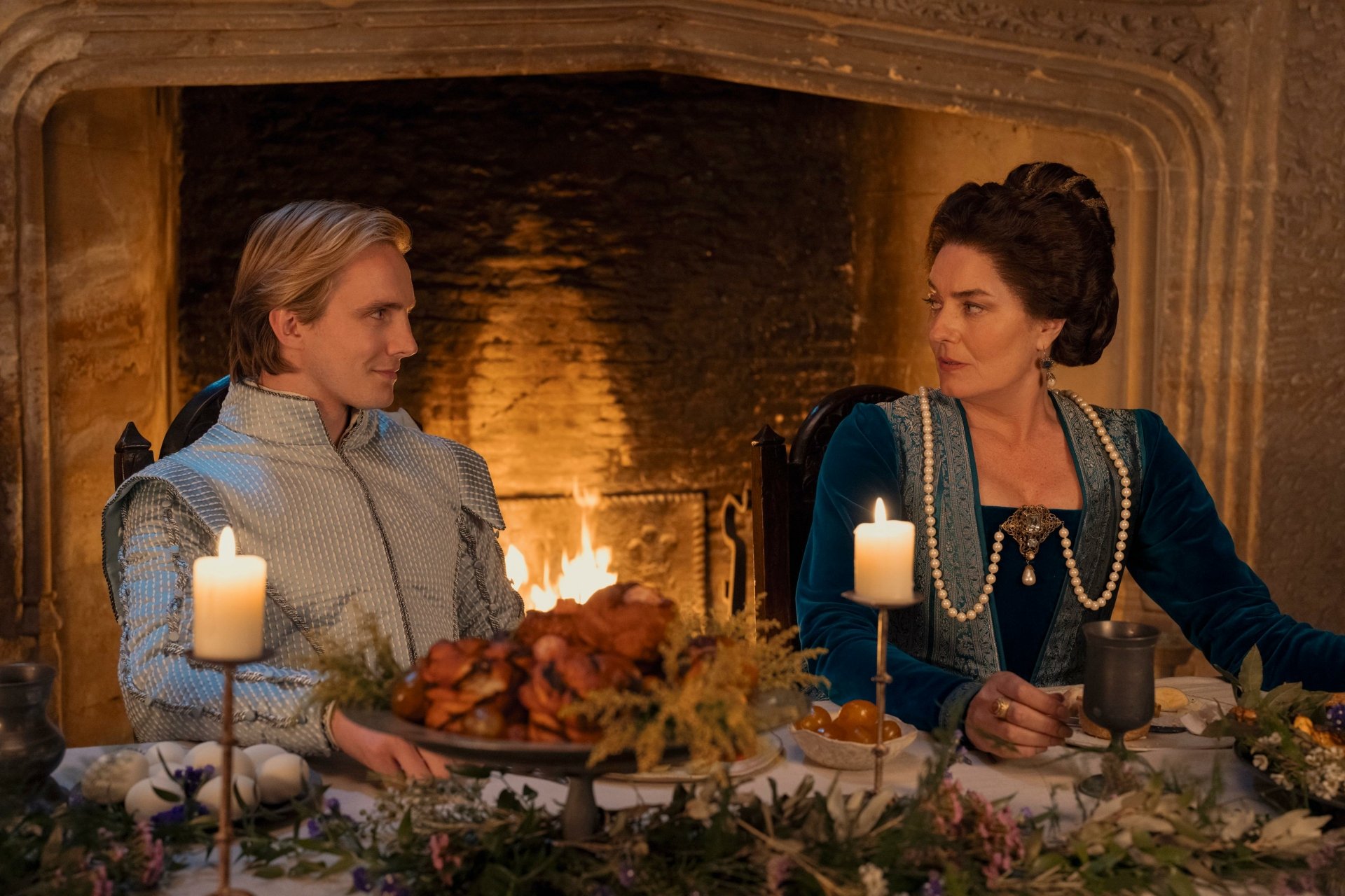 A scene from the movie My Lady Jane featuring two characters in period costumes, sitting at a decorated dinner table with candles, in front of a lit fireplace. HD desktop wallpaper and background.