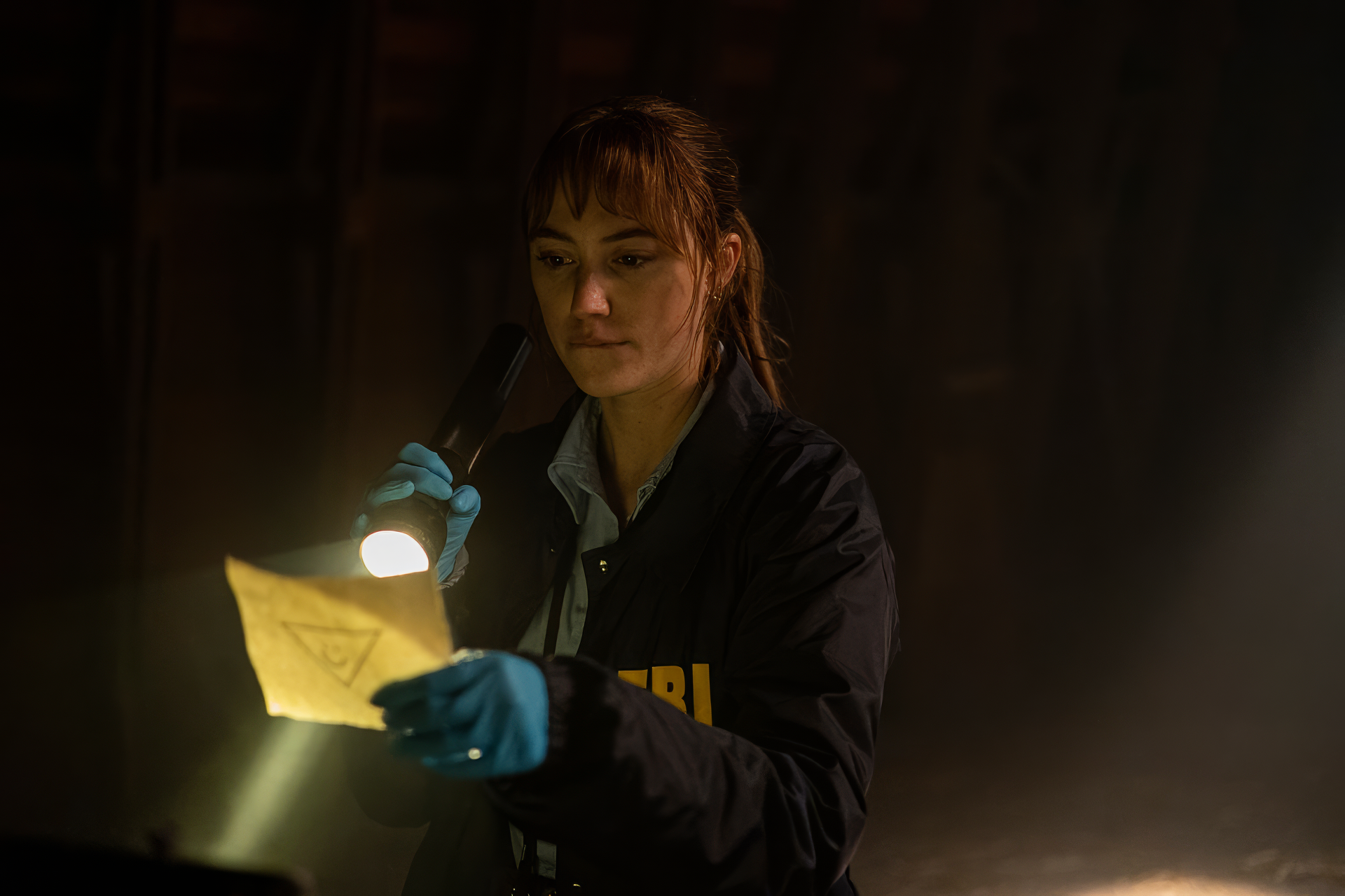 A scene from the movie Longlegs shows an investigator examining a piece of evidence with a flashlight in a dimly lit room. This HD wallpaper serves as a captivating background for desktops.