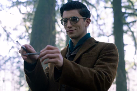 HD desktop wallpaper from the TV show Dead Boy Detectives featuring a young man in a brown jacket and glasses, standing in a forest holding a pen.