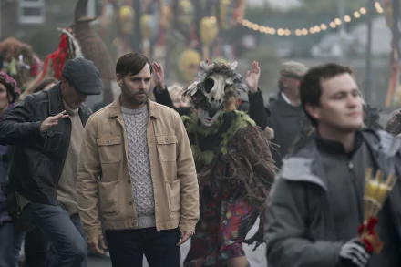 HD desktop wallpaper from the TV show Bodkin featuring a man in a beige jacket amidst a festive, chaotic street scene with masked and costumed characters.