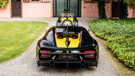 HD wallpaper featuring a black and yellow Bugatti Chiron Super Sport 55 parked on a gravel path in front of a building with greenery. Another classic car is visible ahead of it.