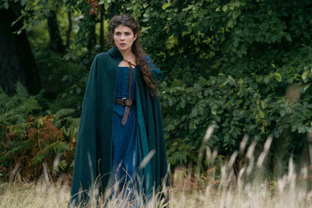 A scene from My Lady Jane movie featuring a woman in a green cloak standing in a lush, wooded area. This image serves as a HD desktop wallpaper and background.