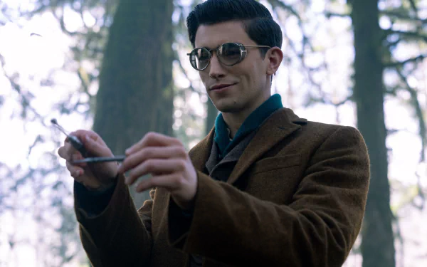 HD desktop wallpaper from the TV show Dead Boy Detectives featuring a young man in a brown jacket and glasses, standing in a forest holding a pen.