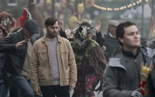 HD desktop wallpaper from the TV show Bodkin featuring a man in a beige jacket amidst a festive, chaotic street scene with masked and costumed characters.