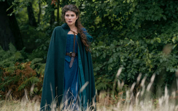 A scene from My Lady Jane movie featuring a woman in a green cloak standing in a lush, wooded area. This image serves as a HD desktop wallpaper and background.